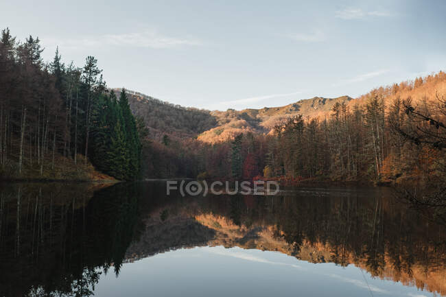 Paesaggio paesaggistico con foresta verde e gialla e colline riflesse nella calma acqua scura del bellissimo lago nella giornata di sole — Foto stock