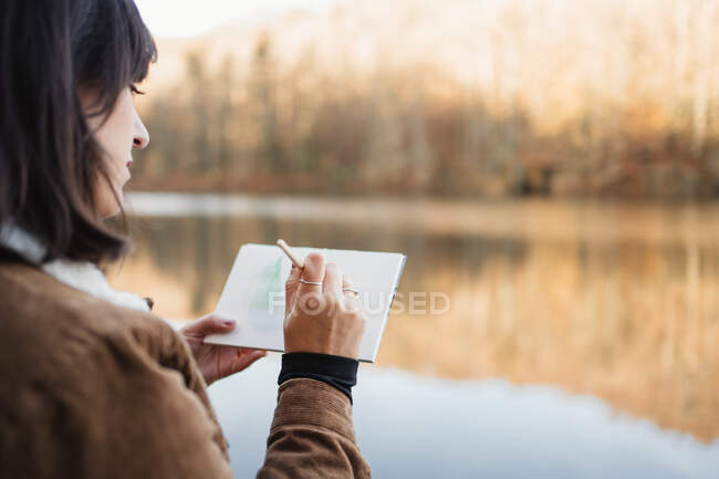 Mulher em pé à beira do lago e desenho em caderno — Fotografia de Stock
