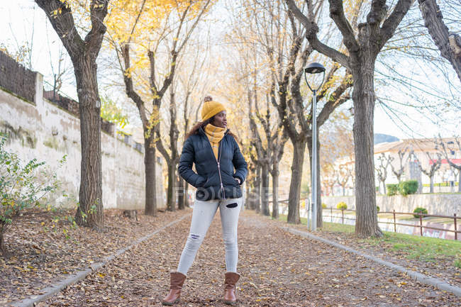 Charming fashion African American woman in yellow hat and warm jacket with hands in pocket on road with autumn leaves in park — Stock Photo