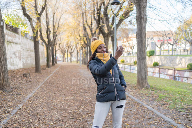 Cheerful fashion African American woman in yellow hat and warm jacket taking selfie on smartphone on road with autumn leaves in park — Stock Photo