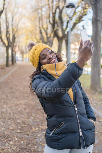 Афроамериканка в жёлтой шляпе и тёплой куртке делает селфи на смартфоне на дороге с осенними листьями в парке — стоковое фото