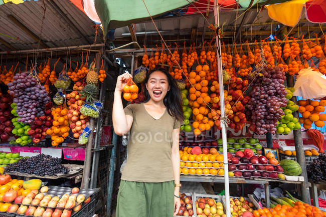 Excitada turista asiática em roupas casuais rindo enquanto segurava o bolso com tangerinas no colorido mercado ao ar livre no Sri Lanka — Fotografia de Stock