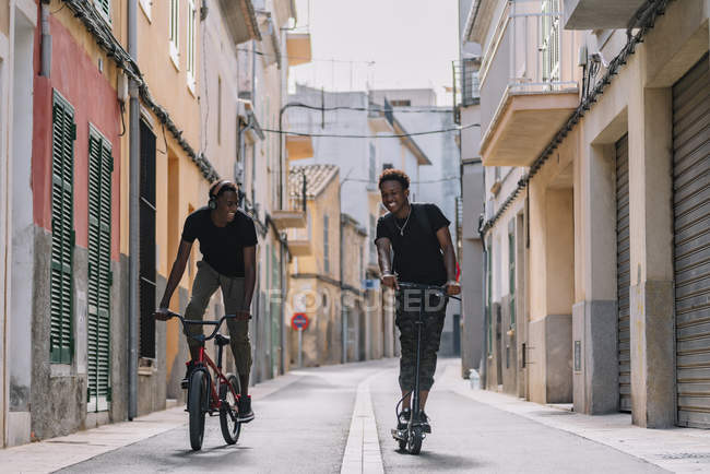 Cheerful young African American man riding electric scooter while black male friend is driving bicycle in street — Stock Photo