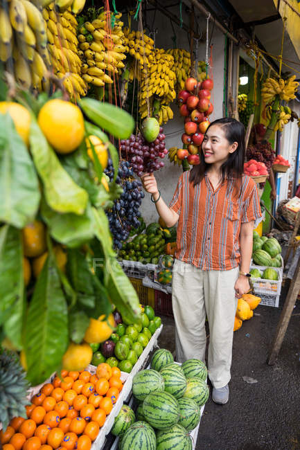 Soddisfatto asiatico riposo signora in casual usura degustazione uva e sorridente all'aperto mercato — Foto stock