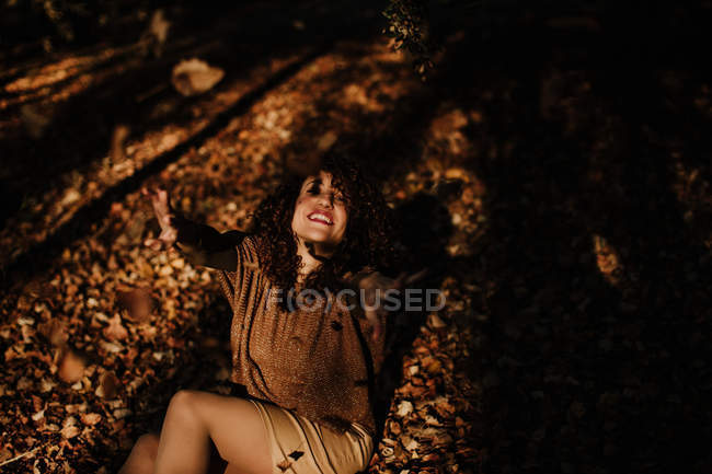Зверху кучеряве волосся задоволена жінка в повсякденному одязі кидає осіннє листя і посміхається в парку — стокове фото