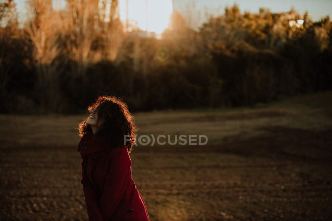 Vista laterale di gioiosa donna rossa in caldo cappotto sorridente e guardando la fotocamera mentre si cammina lungo la foresta autunnale — Foto stock