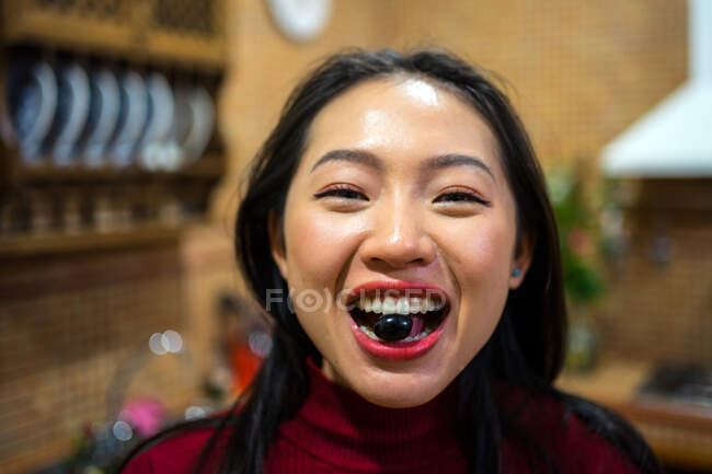 Contenu asiatique femme tenant noir olive dans dents et regarder caméra — Photo de stock