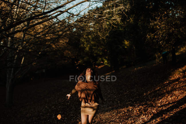 Кудрявая довольная женщина в повседневной одежде танцует в парке полном осенних листьев и улыбается — стоковое фото