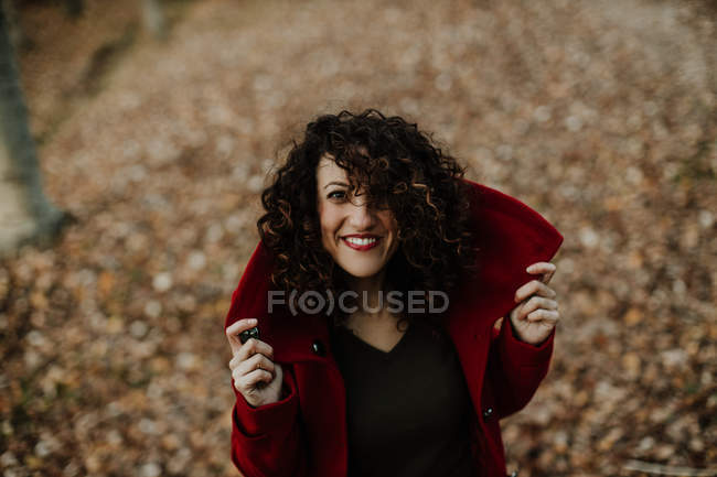 Сверху кудрявая женщина улыбается и смотрит в камеру с золотистыми опавшими листьями на размытом фоне — стоковое фото