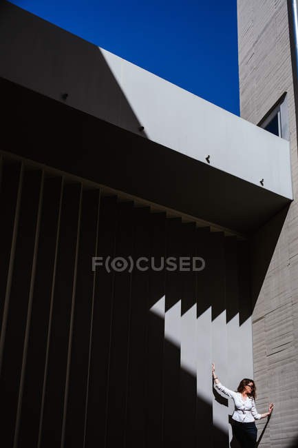 Junge Frau mit trendiger Sonnenbrille und stylischem Outfit hebt an sonnigem Tag die Hand und lehnt an der Wand eines modernen Gebäudes — Stockfoto