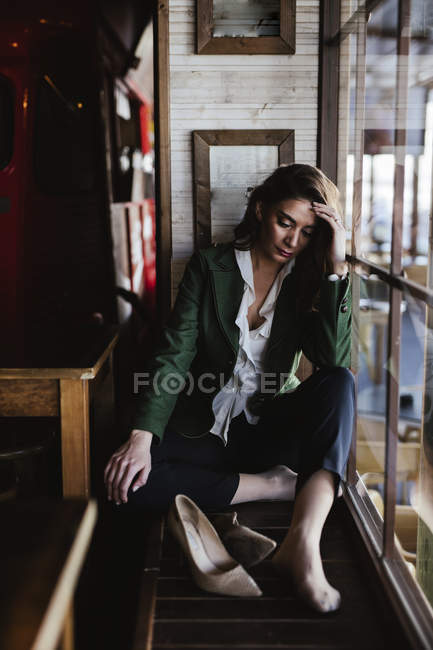 Зверху стомлена модна жінка сидить з схрещеними ногами і дивиться крізь вікно сучасного кафе — стокове фото
