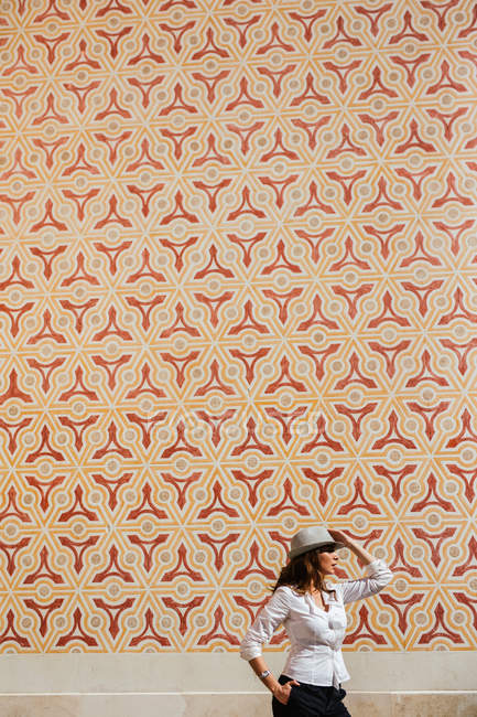 Seitenansicht der jungen Frau lässig stilvolles Outfit hält Hand auf Hut mit ornamentalen Wand auf dem Hintergrund — Stockfoto