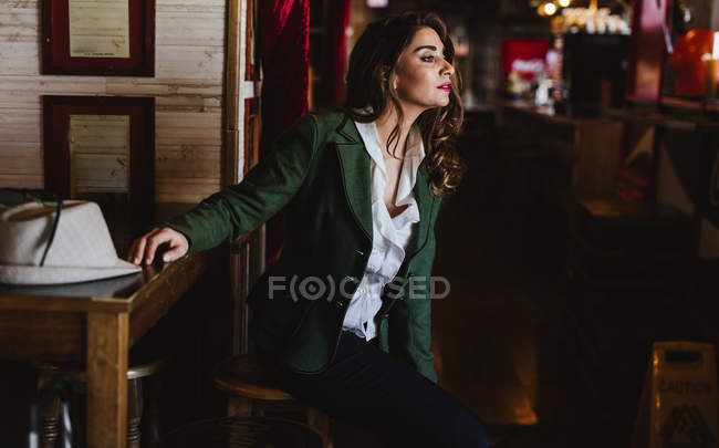 Jeune femme à la mode dans des vêtements à la mode souriant et détournant les yeux tout en se reposant dans un café confortable — Photo de stock