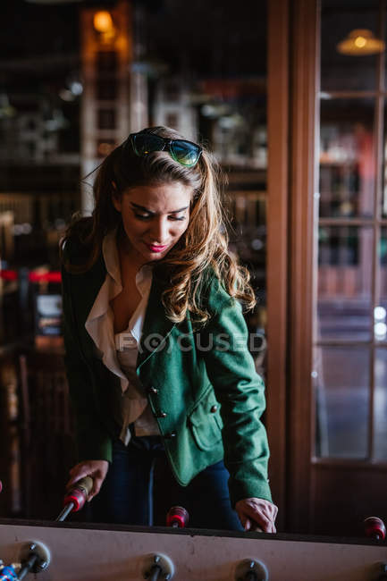 Зверху радісна коричнева волохата жінка в стильному вбранні сміється, граючи в настільний футбол в сучасному кафе — стокове фото