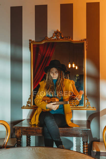 Mujer joven y elegante con rastas con abrigo amarillo y sombrero negro sentado en la vieja mesa de madera de nuevo al espejo y tocando ukelele de guitarra hawaiana en la habitación con muebles antiguos - foto de stock