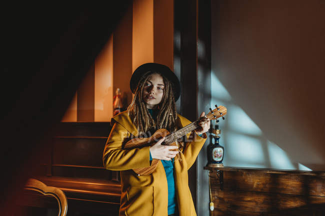 Hipster-Millennial-Frau mit Dreadlocks in gelbem Mantel und schwarzem Hut spielt hawaiianische Gitarrenukulele, während sie im dunklen Vintage-Raum steht — Stockfoto