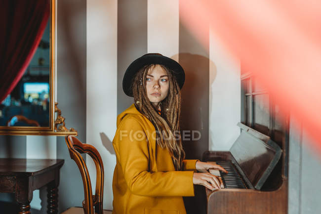 Vista laterale di una donna hipster seria con dreadlocks che indossa un cappotto giallo e un cappello nero che suona il pianoforte mentre è seduta in una stanza in stile retrò — Foto stock