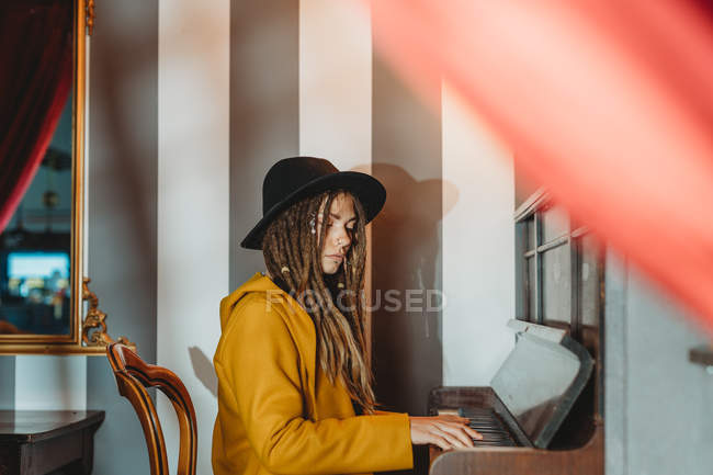 Side view of serious hipster female with dreadlocks wearing yellow coat and black hat playing piano while sitting in retro styled room — Stock Photo