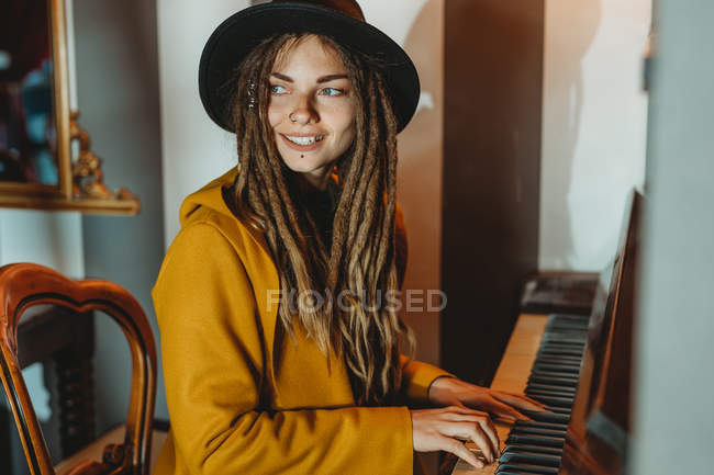 Side view of happy hipster female with dreadlocks wearing yellow coat and black hat playing piano while sitting in retro styled room — Stock Photo