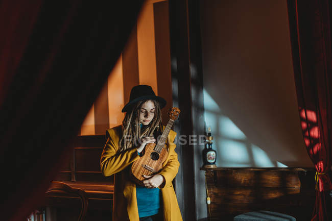 Гіпстерська тисячолітня жінка з дредами в жовтому пальто і чорний капелюх грає на гавайській гітарі укулеле, стоячи в темній старовинній кімнаті. — стокове фото