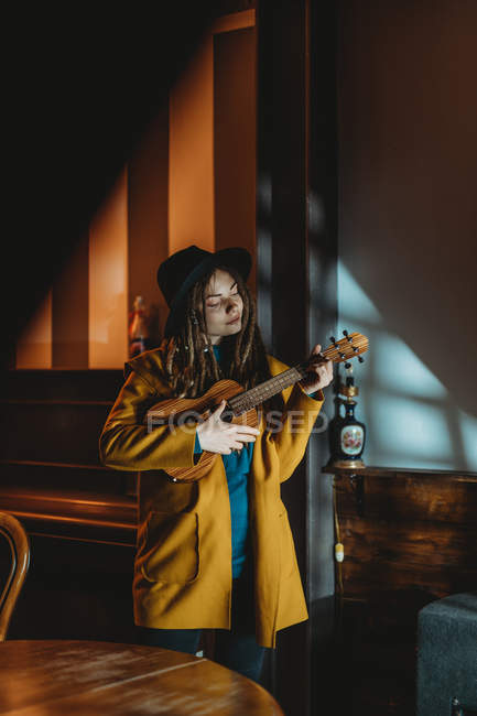 Hipster millennial female with dreadlocks wearing yellow coat and black hat playing Hawaiian guitar ukulele while standing in dark vintage room — Stock Photo