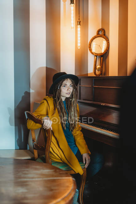 Vista lateral de una hembra hipster seria con rastas con abrigo amarillo y sombrero negro tocando el piano mientras está sentada en una habitación de estilo retro - foto de stock