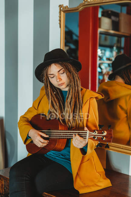 Woman with dreadlocks wearing yellow coat and black hat sitting on old wooden table back to mirror and playing Hawaiian guitar ukulele — Stock Photo