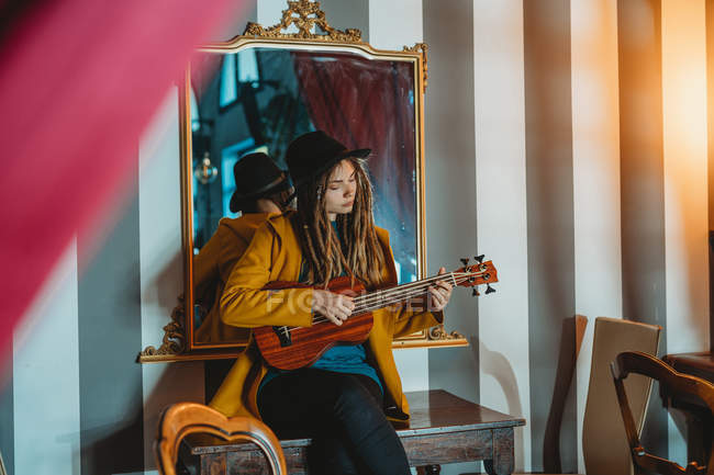 Mujer joven y elegante con rastas con abrigo amarillo y sombrero negro sentado en la vieja mesa de madera de nuevo al espejo y tocando ukelele de guitarra hawaiana - foto de stock