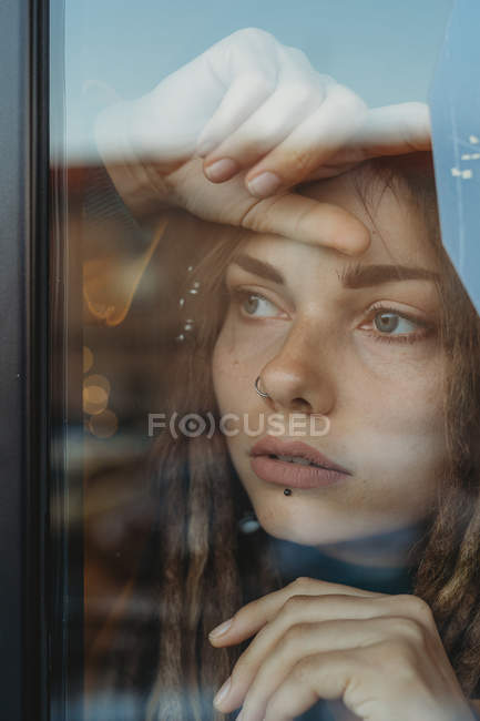 Mujer joven y triste pensativa con rastas apoyadas en el cristal de la ventana y mirando hacia otro lado - foto de stock