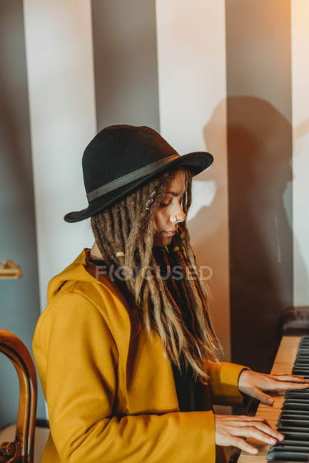 Vista lateral da fêmea hipster grave com dreadlocks vestindo casaco amarelo e chapéu preto tocando piano enquanto sentado em quarto estilo retro — Fotografia de Stock