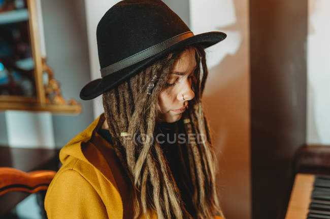 Vista lateral de una hembra hipster seria con rastas con abrigo amarillo y sombrero negro tocando el piano mientras está sentada en una habitación de estilo retro - foto de stock