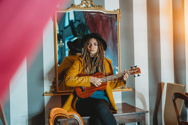 Mujer joven y elegante con rastas con abrigo amarillo y sombrero negro sentado en la vieja mesa de madera de nuevo al espejo y tocando ukelele de guitarra hawaiana - foto de stock