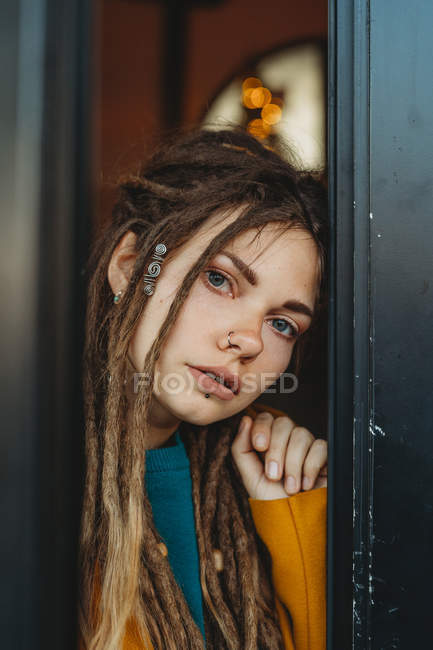 Stylish millennial female with dreadlocks wearing yellow coat over blue sweater looking away thoughtfully while sitting in room — Stock Photo
