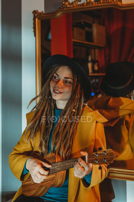Jeune femme élégante avec dreadlocks portant un manteau jaune et un chapeau noir assis sur une vieille table en bois dos au miroir et jouant de la guitare hawaïenne ukulele — Photo de stock