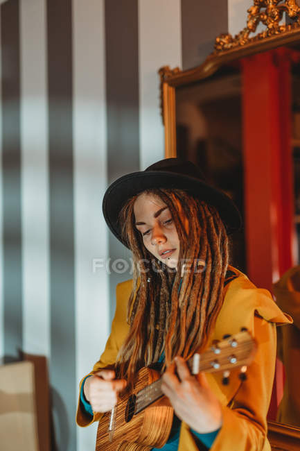 Jeune femme élégante avec dreadlocks portant un manteau jaune et un chapeau noir assis sur une vieille table en bois dos au miroir et jouant de la guitare hawaïenne ukulele — Photo de stock