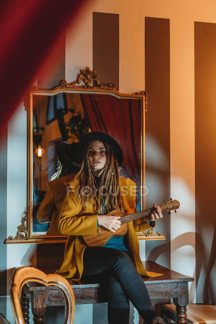 Junge stylische Frau mit Dreadlocks in gelbem Mantel und schwarzem Hut, die auf einem alten Holztisch sitzt und hawaiianische Gitarrenukulele spielt — Stockfoto