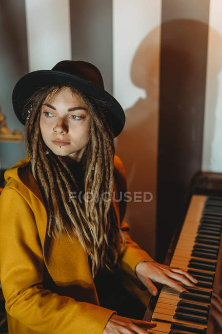 Vue latérale d'une femme hipster sérieuse avec dreadlocks vêtue d'un manteau jaune et d'un chapeau noir jouant du piano assis dans une pièce de style rétro — Photo de stock