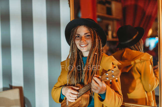 Joven mujer alegre y elegante con rastas con abrigo amarillo y sombrero negro sentado en la vieja mesa de madera de nuevo al espejo y tocando ukelele de guitarra hawaiana - foto de stock
