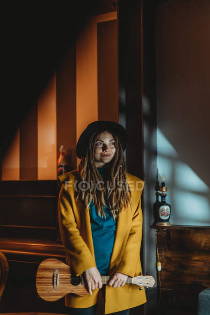 Hipster millennial female with dreadlocks wearing yellow coat and black hat playing Hawaiian guitar ukulele while standing in dark vintage room — Stock Photo