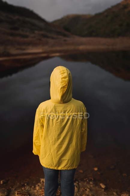 Vista posteriore di persona in impermeabile giallo con cappuccio che copre la testa in piedi vicino al lago nella giornata nuvolosa — Foto stock