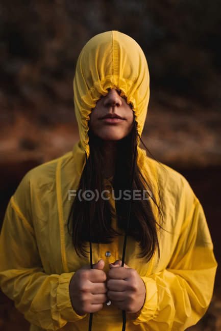 Donna in cappotto pioggia giallo indossa cappuccio che copre il viso in piedi in natura il giorno nuvoloso — Foto stock