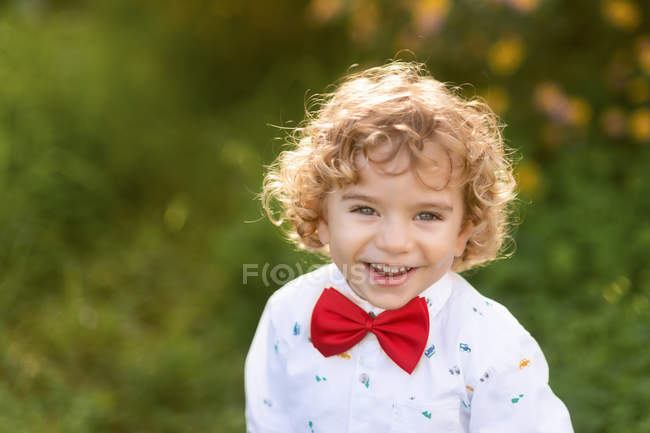 Сверху радостный кудрявый маленький мальчик в рубашке и галстуке-бабочке смеется и смотрит в камеру с зеленой травой на размытом фоне — стоковое фото