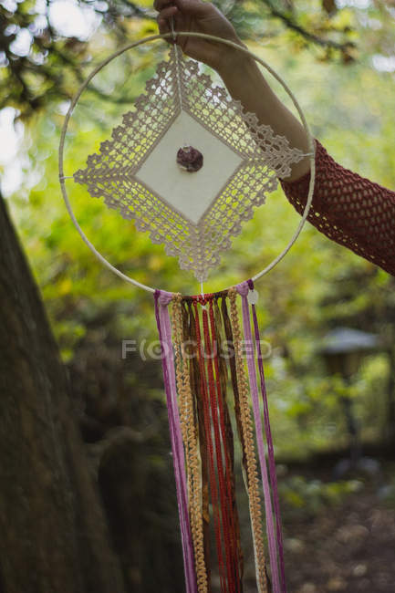 Cropped image of woman in casual wear showing handmade dreamcatchers with long threads looking at amulet spending time in fresh air — Stock Photo