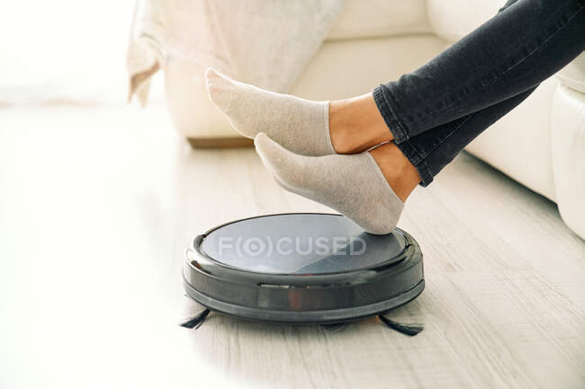 Side view of crop female in jeans and socks sitting on white sofa in room with laminate floor and putting feet on robotic vacuum cleaner — Stock Photo