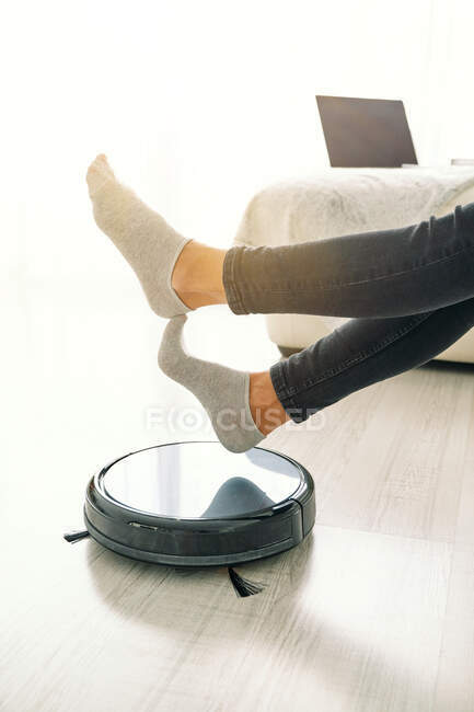 Vue latérale de la culture femelle en jeans et chaussettes assis sur un canapé blanc dans la chambre avec sol stratifié et mettre les pieds sur aspirateur robotique — Photo de stock