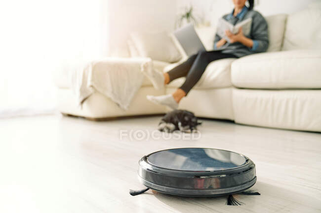 Милый щенок лежит следующий круглый черный роботизированный пылесос в то время как урожай женщина сидит на диване в светлой комнате с ламинатом пол — стоковое фото