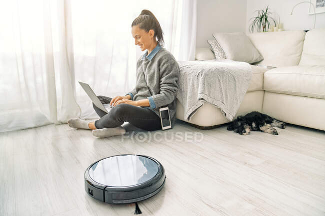 Woman sitting on the floor of a light room working on a computer with cute puppy lying down next round black robotic vacuum cleaner — Stock Photo