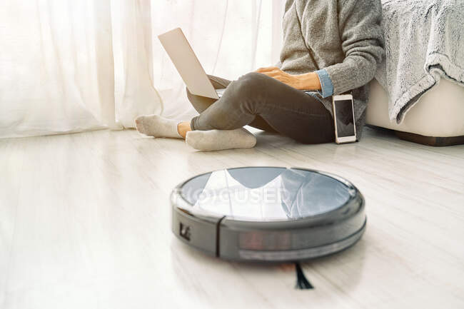 Crop anonymous woman in casual clothe sitting on the floor browsing internet on laptop while robotic vacuum cleaner works — Stock Photo