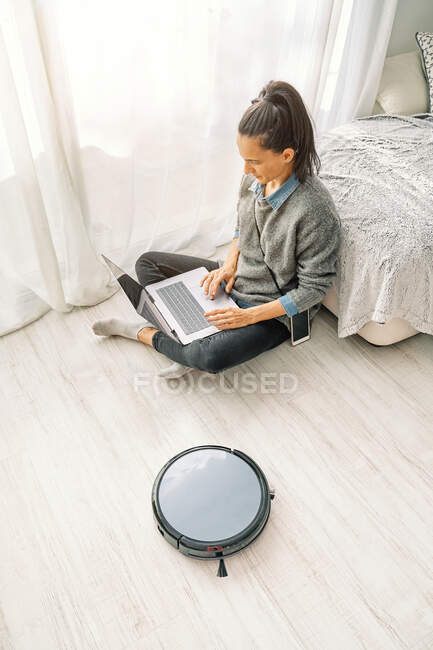 From above woman in casual clothes using remote control for robotic vacuum cleaner while sitting on sofa and enjoying free time browsing internet on laptop — Stock Photo