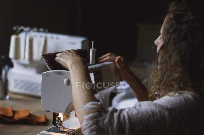 Back view of anonymous crop woman using sewing machine making clothes sitting at table in house — Stock Photo
